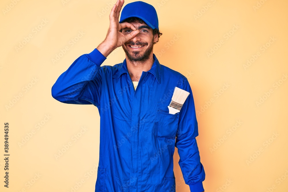 Handsome young man with curly hair and bear wearing builder jumpsuit uniform doing ok gesture with hand smiling, eye looking through fingers with happy face.