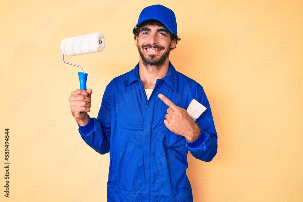 Handsome young man with curly hair and bear wearing builder jumpsuit uniform holding paint roller smiling happy pointing with hand and finger Handsome young man with curly hair and bear wearing builder jumpsuit uniform holding paint roller smiling happy pointing with hand and finger
