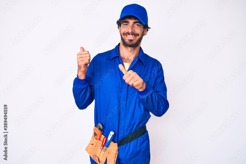 Handsome young man with curly hair and bear weaing handyman uniform pointing to the back behind with hand and thumbs up, smiling confident