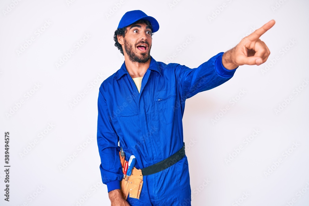 Handsome young man with curly hair and bear weaing handyman uniform pointing with finger surprised ahead, open mouth amazed expression, something on the front Handsome young man with curly hair and bear weaing handyman uniform pointing with finger surprised ahead, open mouth amazed expression, something on the front