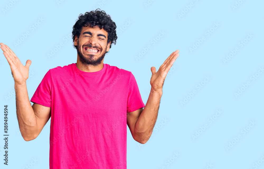 Handsome young man with curly hair and bear wearing casual pink tshirt celebrating mad and crazy for success with arms raised and closed eyes screaming excited. winner concept Handsome young man with curly hair and bear wearing casual pink tshirt celebrating mad and crazy for success with arms raised and closed eyes screaming excited. winner concept