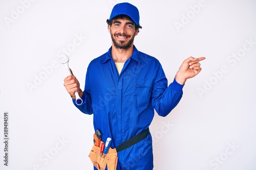 Handsome young man with curly hair and bear wearing builder jumpsuit uniform and holding wrench smiling happy pointing with hand and finger to the side