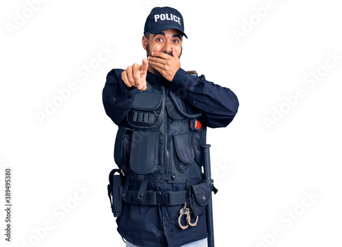 Young hispanic man wearing police uniform laughing at you, pointing finger to the camera with hand over mouth, shame expression