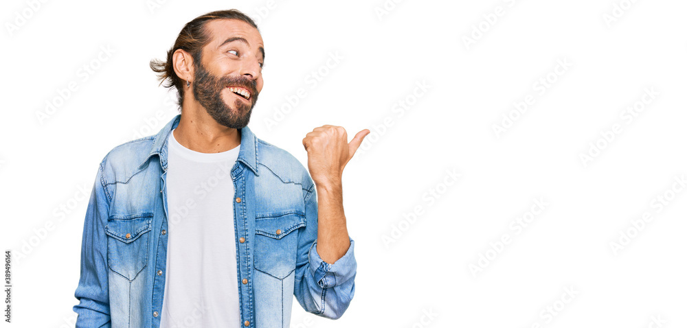 Attractive man with long hair and beard wearing casual denim jacket smiling with happy face looking and pointing to the side with thumb up. Attractive man with long hair and beard wearing casual denim jacket smiling with happy face looking and pointing to the side with thumb up.