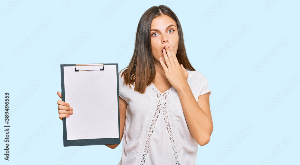 Young caucasian woman holding clipboard with blank space covering mouth with hand, shocked and afraid for mistake. surprised expression Young caucasian woman holding clipboard with blank space covering mouth with hand, shocked and afraid for mistake. surprised expression