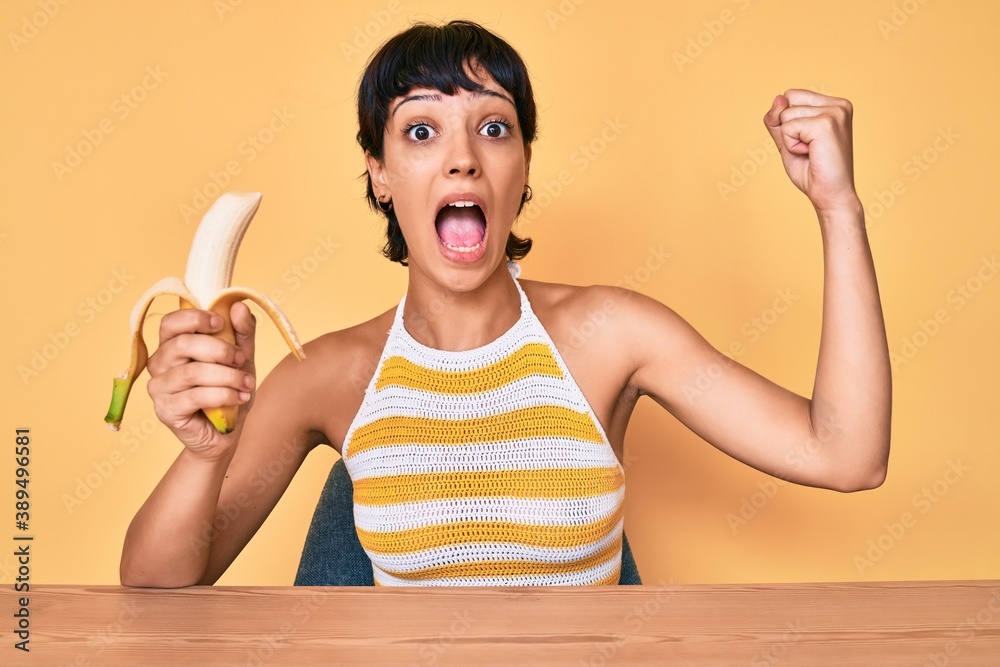Brunette teenager girl eating banana as healthy snack afraid and shocked with surprise and amazed expression, fear and excited face.