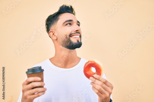 Young arab man smiling happy eating donut and drinking coffee at the city.