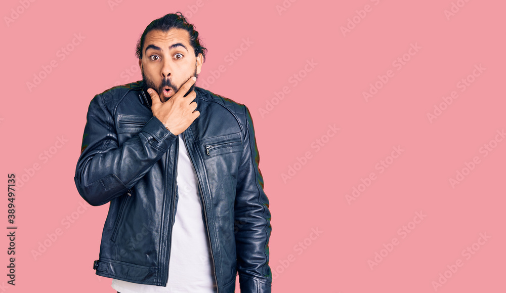 Young arab man wearing casual leather jacket looking fascinated with disbelief, surprise and amazed expression with hands on chin