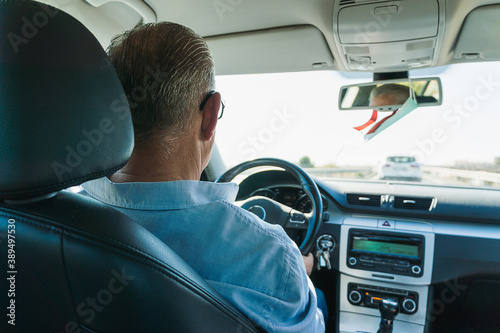 Caucasian male with gray hair and about 50 years old driving a car on the road. The driver is dressed in a blue shirt and wears eye glasses. The car is spacious and stately.