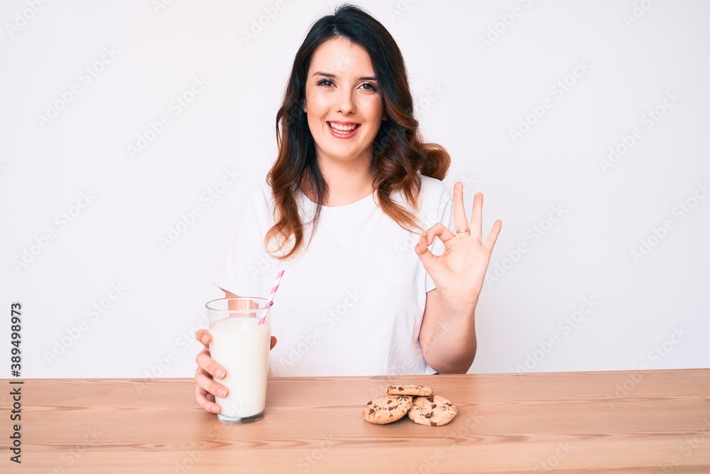 Young beautiful brunette woman drinking a glass of fresh milk with cookies doing ok sign with fingers, smiling friendly gesturing excellent symbol
