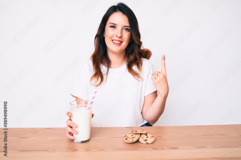 Young beautiful brunette woman drinking a glass of fresh milk with cookies smiling with an idea or question pointing finger with happy face, number one Young beautiful brunette woman drinking a glass of fresh milk with cookies smiling with an idea or question pointing finger with happy face, number one