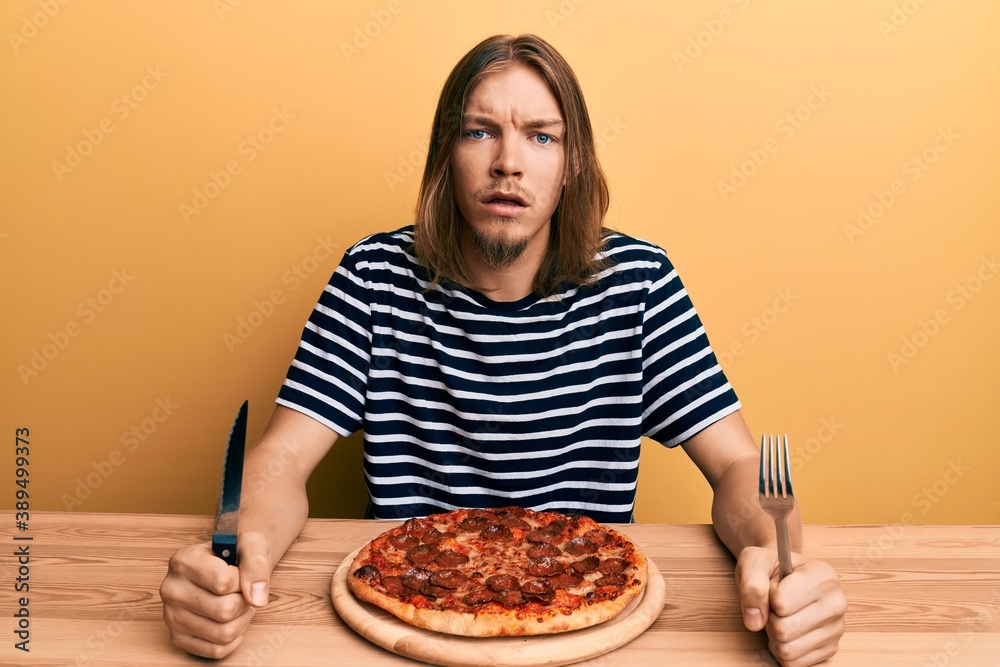Handsome caucasian man with long hair eating tasty pepperoni pizza in shock face, looking skeptical and sarcastic, surprised with open mouth Handsome caucasian man with long hair eating tasty pepperoni pizza in shock face, looking skeptical and sarcastic, surprised with open mouth