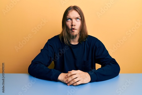 Handsome caucasian man with long hair wearing casual clothes sitting on the table making fish face with lips, crazy and comical gesture. funny expression. © Krakenimages.com