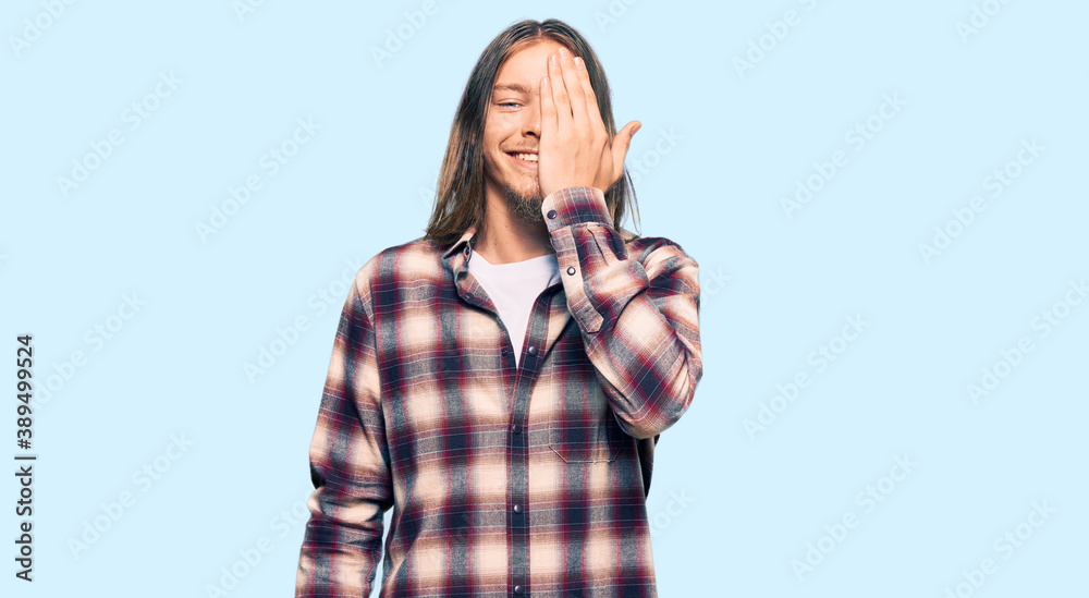 Handsome caucasian man with long hair wearing hipster shirt covering one eye with hand, confident smile on face and surprise emotion.