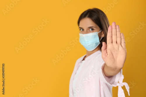 Woman in protective mask showing stop gesture on yellow background, space for text. Prevent spreading of coronavirus © New Africa