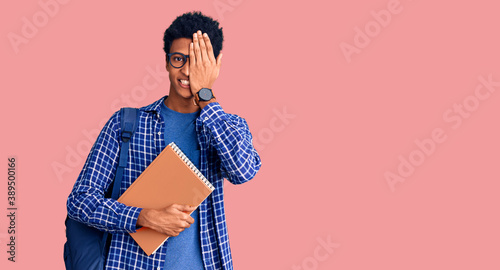 Young african american man wearing student backpack holding book covering one eye with hand, confident smile on face and surprise emotion.