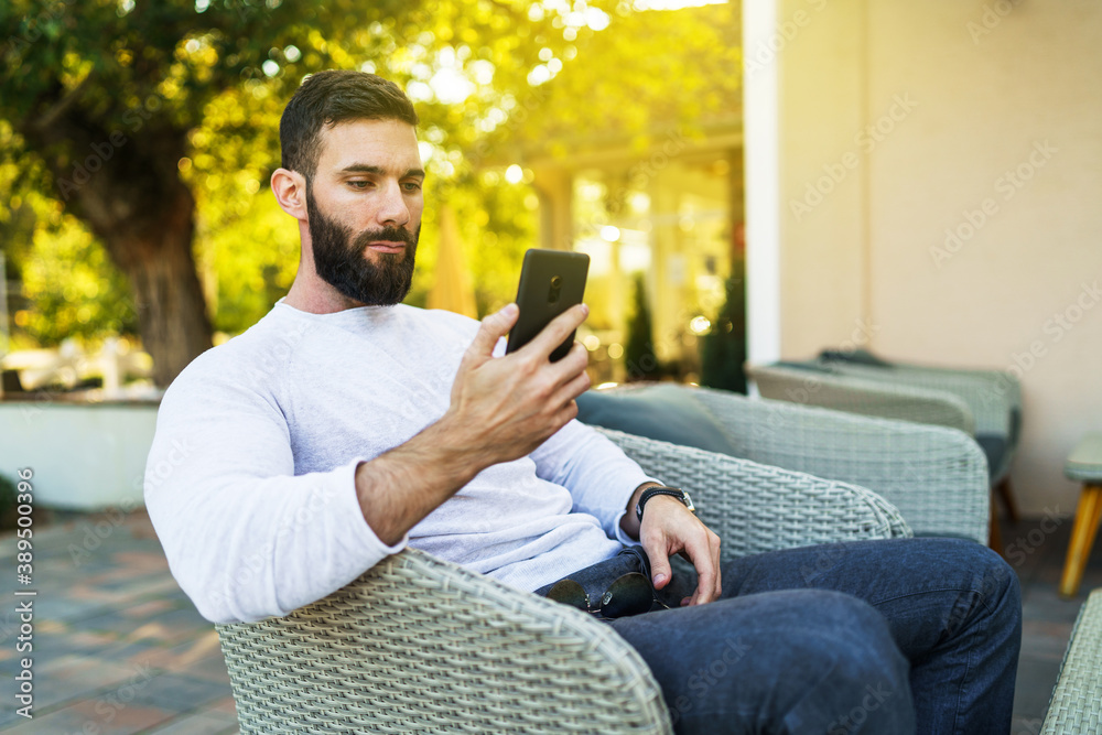 Side view portrait of adult modern caucasian man with beard sitting outdoor serious using mobile phone for video call at cafe in autumn day - leisure concept copy space