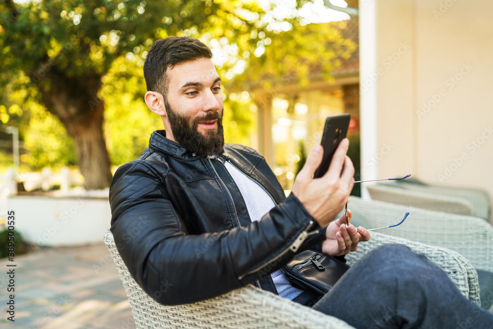 Side view portrait of adult modern caucasian man with beard sitting outdoor surprised and happy using mobile phone for video call at cafe in sunny autumn day - leisure concept copy space Side view portrait of adult modern caucasian man with beard sitting outdoor surprised and happy using mobile phone for video call at cafe in sunny autumn day - leisure concept copy space