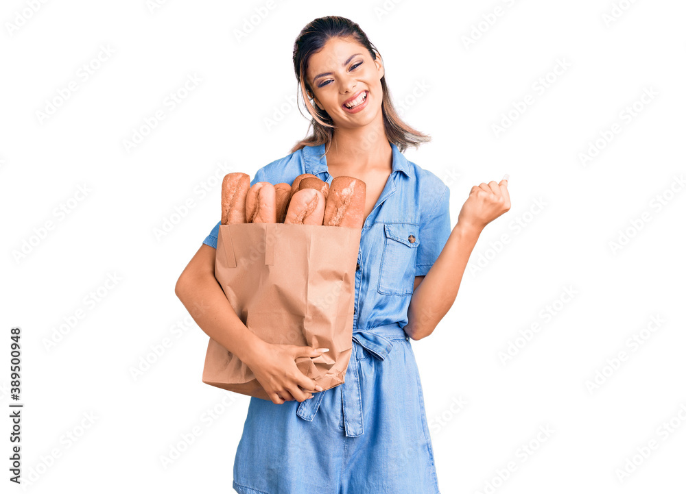 Young beautiful woman holding paper bag with bread screaming proud, celebrating victory and success very excited with raised arms Young beautiful woman holding paper bag with bread screaming proud, celebrating victory and success very excited with raised arms