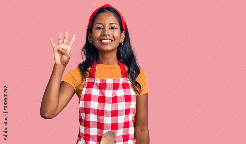 Young indian girl wearing professional baker apron showing and pointing up with fingers number four while smiling confident and happy. Young indian girl wearing professional baker apron showing and pointing up with fingers number four while smiling confident and happy.