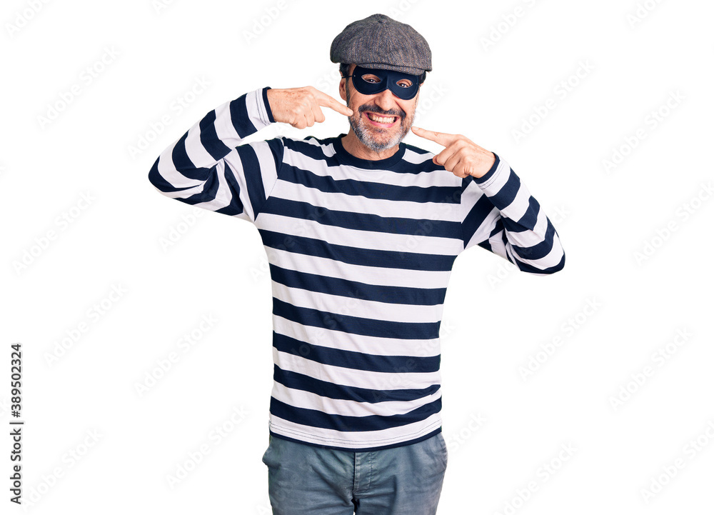 Middle age handsome man wearing burglar mask smiling cheerful showing and pointing with fingers teeth and mouth. dental health concept. Middle age handsome man wearing burglar mask smiling cheerful showing and pointing with fingers teeth and mouth. dental health concept.