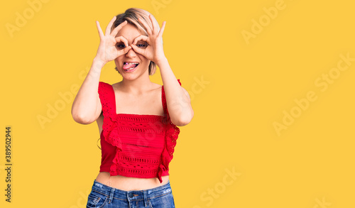 Young beautiful blonde woman wearing casual clothes doing ok gesture like binoculars sticking tongue out, eyes looking through fingers. crazy expression.