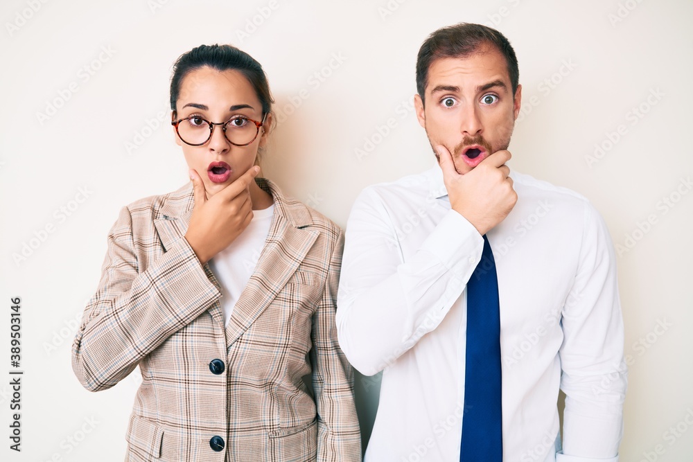Beautiful couple wearing business clothes looking fascinated with disbelief, surprise and amazed expression with hands on chin Beautiful couple wearing business clothes looking fascinated with disbelief, surprise and amazed expression with hands on chin