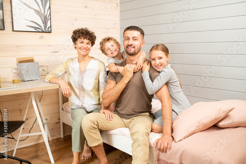 Happy young family of four in casualwear sitting on bed in front of camera