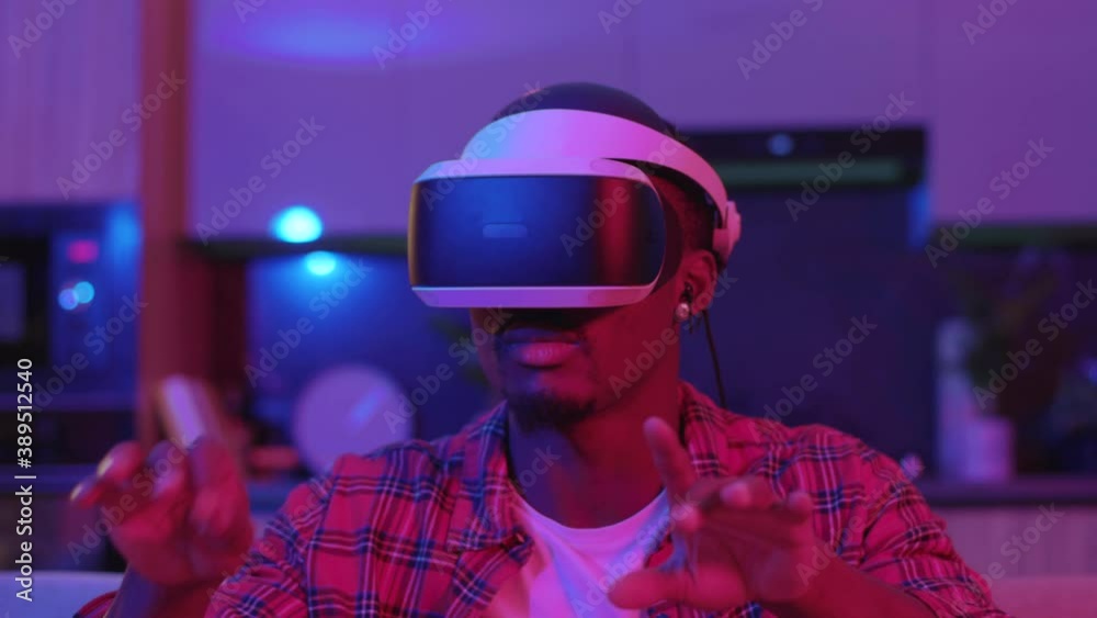At home gaming person young man wearing VR glasses entertaining augmented reality playing virtual simulation interactive games. Neon light. Modern lifestyle. At home gaming person young man wearing VR glasses entertaining augmented reality playing virtual simulation interactive games. Neon light. Modern lifestyle.