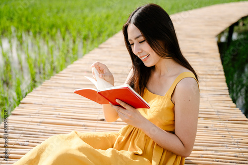 Asian traveler woman in yellow dress reading red book on wooden bridge across rice field. photo