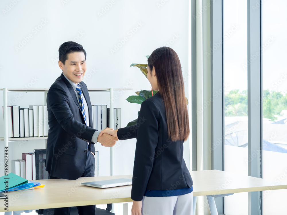 Asian businessman and businesswoman shaking hands in office. Smiling manager standing at his desk in his office handshaking with young female, job applicant after job interview.