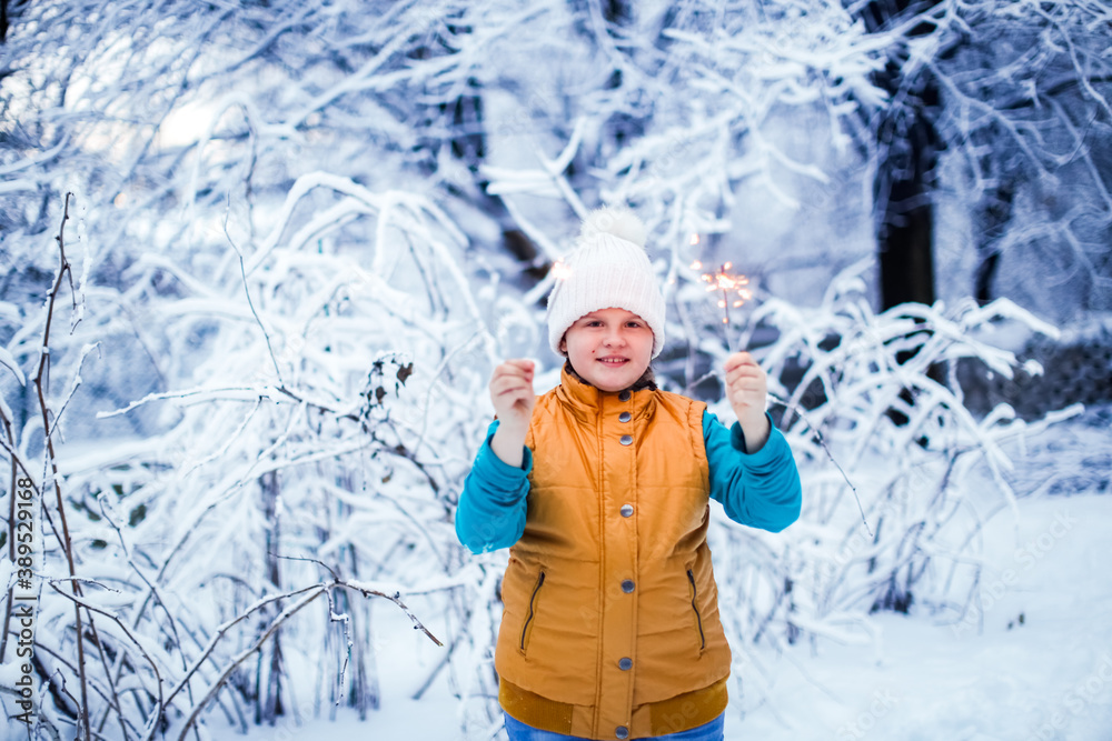 Fototapeta premium Kid girl in yellow winter vest and white knitted hat on a snow scooter with sparklers with the first snow, snowfall and a walk in the park in the backyard. Frost and snowy weather.