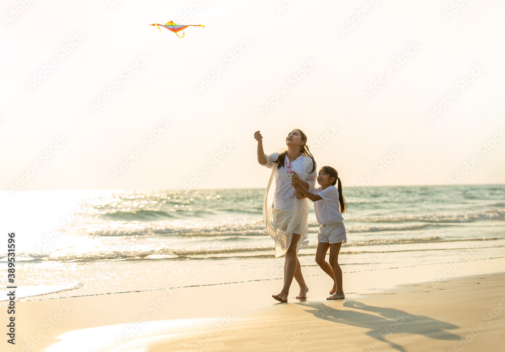 Happy Asian family single mother with little daughter playing kite together on the beach at summer sunset. Beautiful mother and cute child girl relax and having fun in summer holiday vacation.