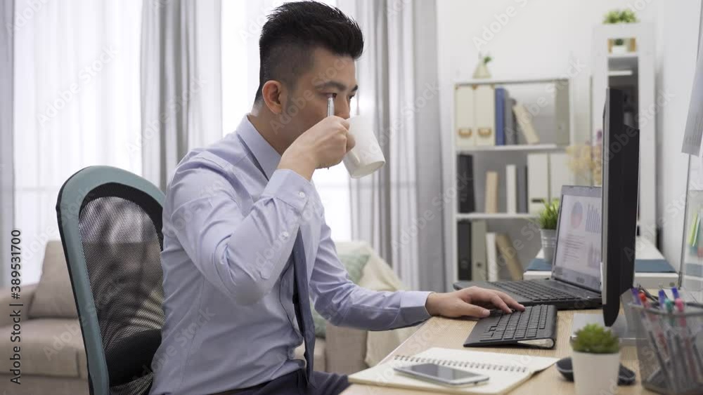 asian male employee drinking coffee while looking at analytical graphs, typing report on computer and taking notes with pen asian male employee drinking coffee while looking at analytical graphs, typing report on computer and taking notes with pen