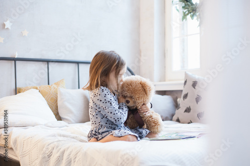 The room is decorated for the new year, a girl is sitting on the bed with a bear, a plush toy is playing school, reading a book, leafing through, a preschooler