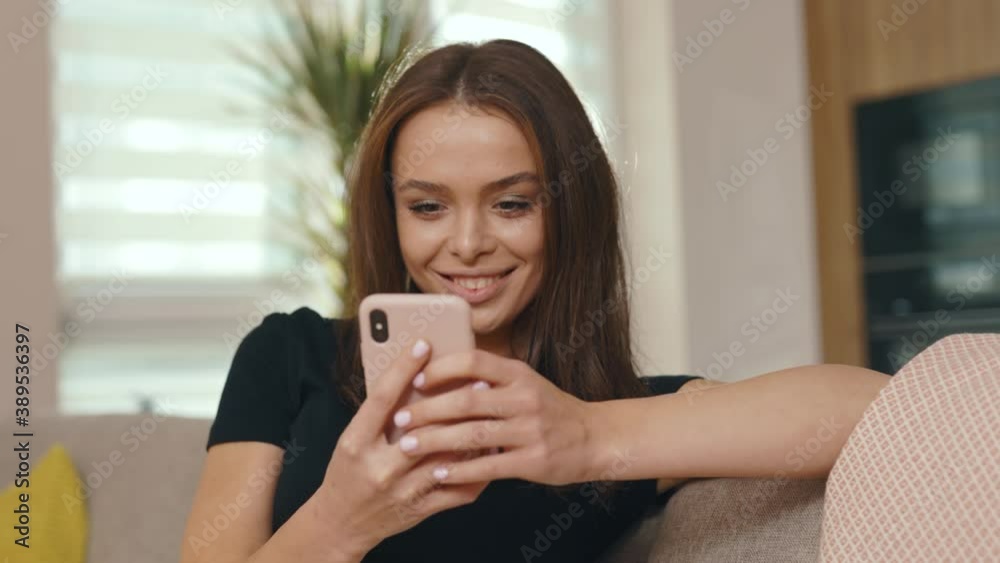 Face young woman sitting on sofa and using smartphone in modern room. Feel happy smiling. Look around at sunny room. Internet social media connection. Slow motion Face young woman sitting on sofa and using smartphone in modern room. Feel happy smiling. Look around at sunny room. Internet social media connection. Slow motion