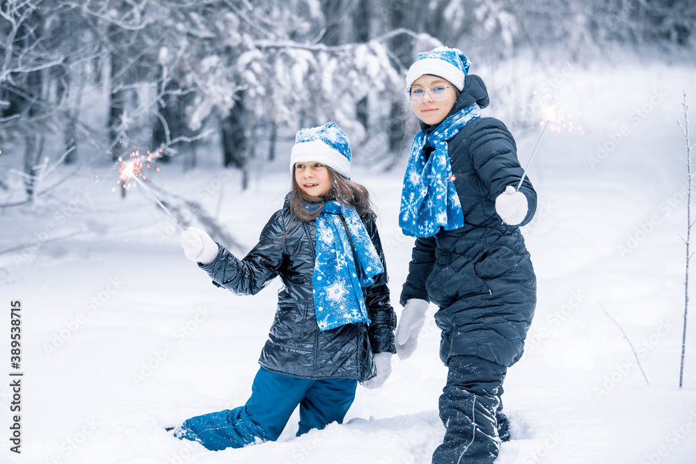 Smiling young girls with sparklers in hands. Happy winter time in the forest. The girls are in a dark coat,  blue  Santa Claus hat, scarf and mittens. Christmas, new year. Festive garland lights.