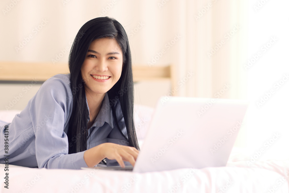 Young and beautiful Asian woman laying on a bed in bedroom with friendly smile and looking to camera and using notebook laptop computer with happiness manner. Concept for modern woman lifestyle