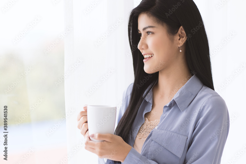 Young and beautiful Asian woman holding white ceramic coffee cup with relax and easy gesture while looking far to outside with soft curtain in background. Idea for happy in the morning time Young and beautiful Asian woman holding white ceramic coffee cup with relax and easy gesture while looking far to outside with soft curtain in background. Idea for happy in the morning time