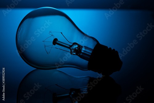 An incandescent light bulb lies on reflective glass, against a blue background, in a dark key.
