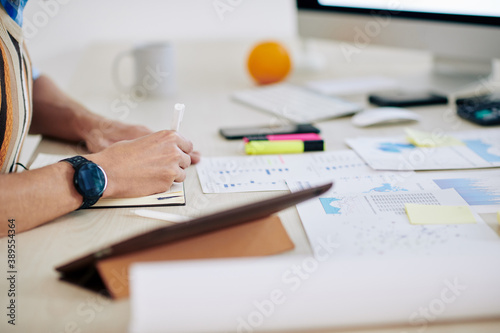 Hands of businessman checking financial charts and writing important figures in planner