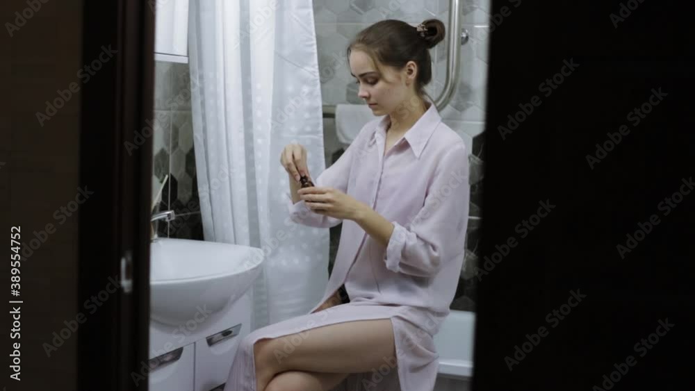 Side view of pretty young woman sitting in bathroom and applying facial serum on hand and face while doing morning skin care routine and looking in mirror Side view of pretty young woman sitting in bathroom and applying facial serum on hand and face while doing morning skin care routine and looking in mirror