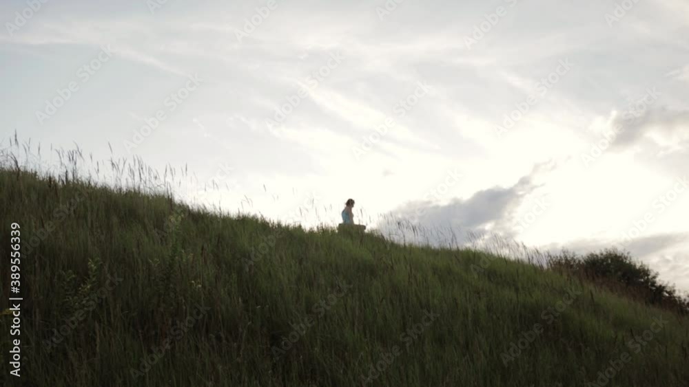 Slim woman with yoga mat walking along grassy hill in summer at sunset Slim woman with yoga mat walking along grassy hill in summer at sunset