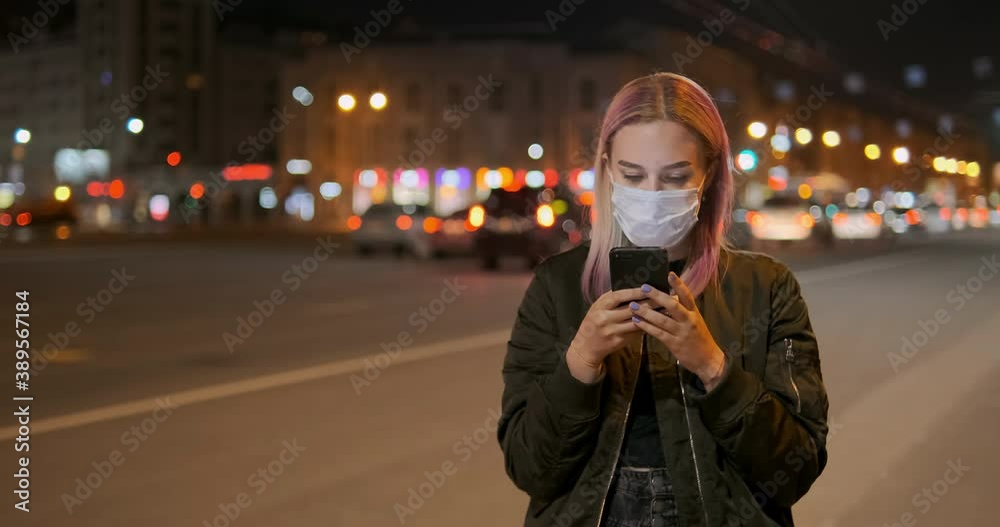 Concentrated woman wearing medical mask looks in smartphone reading sms against evening driving car lights slow motion zoom out Concentrated woman wearing medical mask looks in smartphone reading sms against evening driving car lights slow motion zoom out