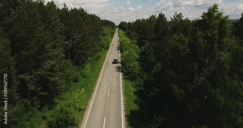 One black SUV car driving alone / travel on empty straight asphalt road freeway through dense forest corridor at summer sunny day. Aerial drone wide view.