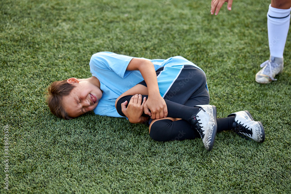 little caucasian boy fell to his knee, ache in joints during football game outdoors, injury during sport competition little caucasian boy fell to his knee, ache in joints during football game outdoors, injury during sport competition