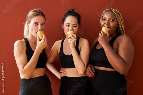 three funny women friends of diverse appearance standing isolated over red background eating ice cream  multi-ethnic group of females enjoy sweet ice cream  smile