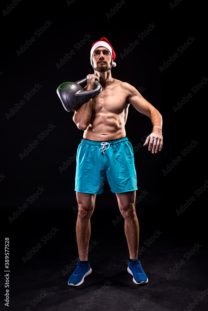 Athletic man in a Santa hat trains with a kettlebell on a black background.