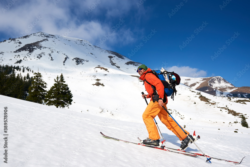 Skier tourist backpacker, happy smiling bearded man hiking on skis up hill on background of blue sky and beautiful mountains ridges. Winter vacations, active lifestyle, skiing and trekking concept.