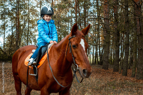 Little girl on an adult brown horse on the background of nature. Jockey, epodrome, horseback riding.